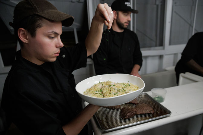 Cook in a commercial kitchen sprinkling fresh herbs over a bowl of creamy risotto, with grilled steak on a wire rack and prep containers on the counter