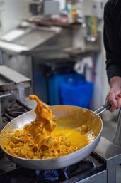 Hand stirring tortellini pasta in a creamy orange cheese sauce in a skillet over a gas stove in a commercial kitchen