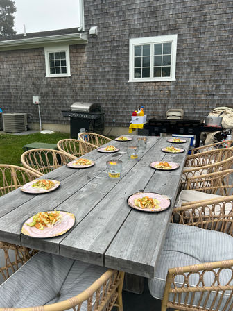 Backyard patio at a shingle-sided house with a long weathered wooden dining table set for an outdoor meal — pink plates of salad at each rattan cushioned chair, candles on the table, and a grill and condiments in the background.