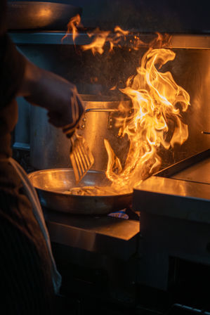 Large flame leaping from a sauté pan as a cook flips food with a spatula on a commercial restaurant stove.