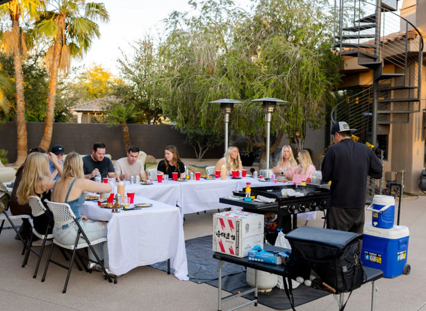 Friends enjoying an outdoor backyard patio dinner at sunset: long white tables with red cups, a chef at a portable grill, palm trees and a spiral staircase.