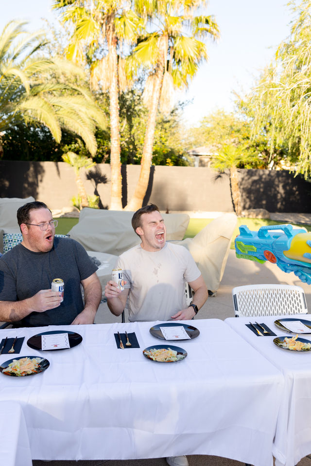 Two men at a white-clothed backyard patio table in the sun, holding beer cans and reacting as a colorful water gun sprays from the right; palm trees and salad plates visible.