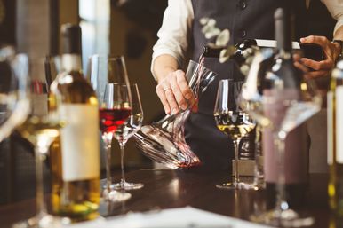Sommelier pouring red wine into a glass decanter at a wine-tasting table surrounded by bottles and filled wine glasses