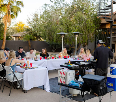 Friends enjoying an outdoor backyard dinner around long white-clothed tables with red cups while a cook grills on a portable griddle on a palm-lined patio with patio heaters and a spiral staircase