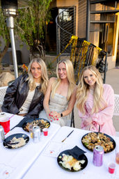 Three smiling blonde friends at an outdoor patio dinner, seated at a white-tablecloth table with plates and drinks, gold-and-black streamers and a spiral staircase in the background, casual evening vibe.