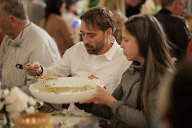 Two diners sharing a large white bowl of creamy risotto at a lively communal dinner table, man scooping a serving with a spoon.