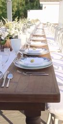 Sunlit al fresco long wooden table set for a reception — farmhouse tablescape with white plates, glassware, folded white napkins topped with small yellow accents, silverware and a lush white floral runner.