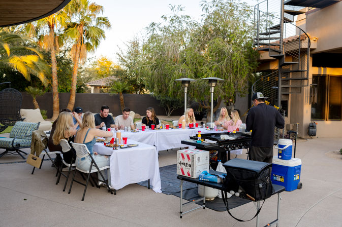 Group enjoying a backyard patio dinner at sunset under palm trees — long white-table setup with red cups, a chef at a portable grill, patio heaters and a spiral outdoor staircase nearby.