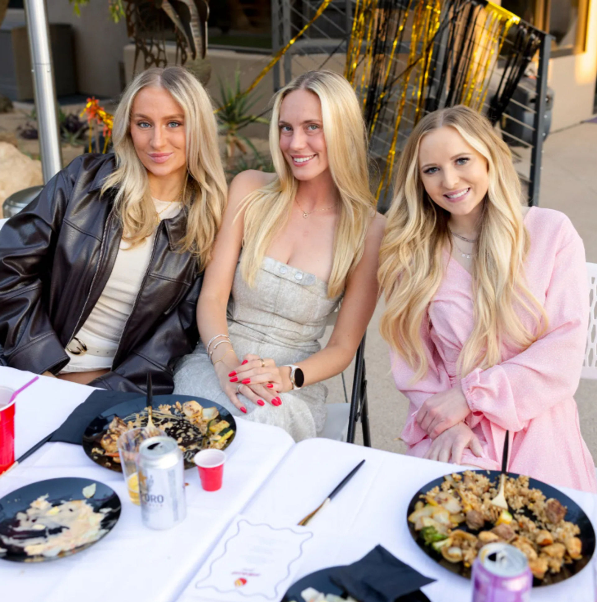 Three smiling women with long blonde hair seated at an outdoor patio table, enjoying a casual celebration with plates of food, drinks, and black-and-gold streamers