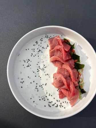 Overhead shot of vibrant pink tuna sashimi slices on seaweed in a white bowl, served with creamy white sesame sauce dotted with black sesame seeds and thin chili slices on a dark tabletop