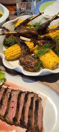 Close-up of charred grilled lamb chops and corn on the cob with dill garnish, served with sliced medium-rare steak on a white plate