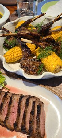 Close-up of charred grilled lamb chops and corn on the cob with dill garnish, served with sliced medium-rare steak on a white plate