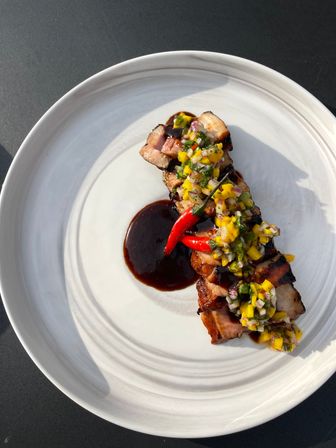 Overhead shot of grilled pork belly slices topped with vibrant mango-chili salsa and two red chilies, plated with a glossy dark glaze on a white ceramic plate in sunlight.