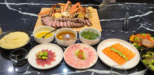 Gourmet seafood and steak spread on a black marble countertop: wooden board with sliced roast beef and crab legs with lemon, bowls of sauces and chimichurri, plates of salmon, tuna and white-fish sashimi, mashed potatoes and mixed salads.
