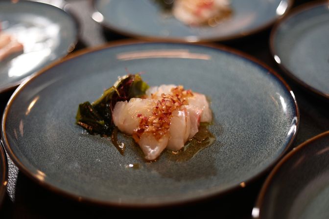 Fine-dining sashimi appetizer: delicate raw fish slices topped with chopped shallots and vinaigrette, served with seaweed on a blue ceramic plate.