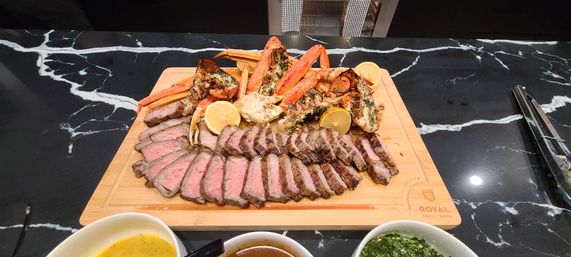 Surf-and-turf platter of sliced medium-rare steak with grilled lobster tails and crab legs on a wooden cutting board, lemon wedges and dipping sauces on a black marble countertop