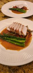 Gourmet seared pork loin, thinly sliced and served with vibrant asparagus and sautéed bell peppers in a glossy brown pan sauce on a white dinner plate over a granite countertop.