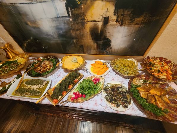 Decorative indoor buffet table with assorted hot dishes and salads — rice, grilled fish, roasted vegetables, fresh herbs and radishes — served on ornate gold and ceramic platters beneath an abstract wall painting.