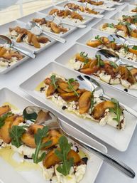 Rows of plated appetizers: creamy burrata topped with grilled peach slices, arugula, olive oil and balsamic drizzle on white rectangular dishes with spoons — beachside catering display.