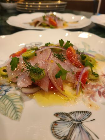 Vibrant raw fish crudo drizzled with olive oil, topped with red onion, cherry tomatoes, sliced green olives, dill and parsley on a floral butterfly china plate.