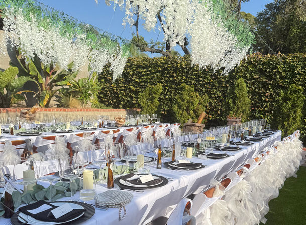 Sunlit outdoor garden banquet: long white-draped tables with black chargers, stacked wine glasses, candles and place cards, ruffled chair sashes and hanging white floral garlands against a green hedge — elegant reception setup.
