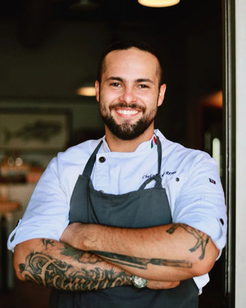 Smiling chef in white coat and gray apron with tattooed forearms, arms crossed at a restaurant doorway under warm pendant lights.