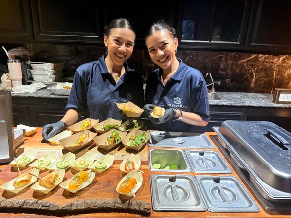 Two smiling catering staff serving bite-sized appetizers in paper boats on a wooden counter at an indoor buffet station