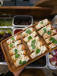 Wooden board of crispy mini flautas — bite-sized rolled tacos drizzled with crema, sprinkled with crumbled cheese and cilantro — presented as an appetizer platter over a food-prep counter with grapes and charcuterie trays nearby.