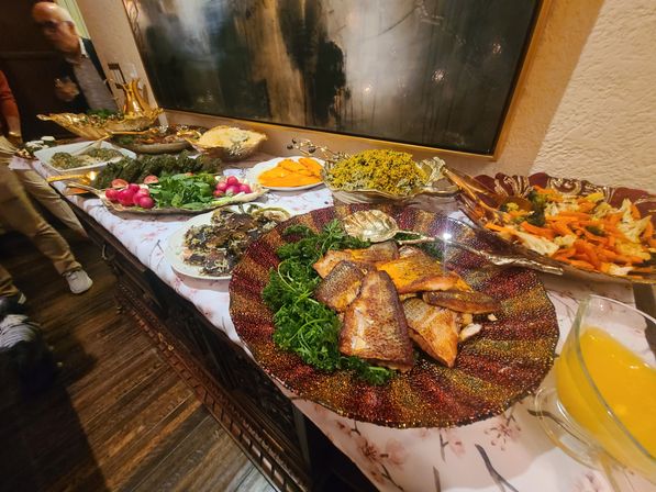 Indoor buffet table with a glittery platter of grilled fish over greens, bowls of roasted vegetables, salads, radishes and a pitcher of orange juice