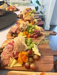 Colorful charcuterie boards lined up on a kitchen counter with green and red grapes, sliced cheeses, prosciutto and salami, assorted crackers, olives and dried apricots on wooden serving boards.