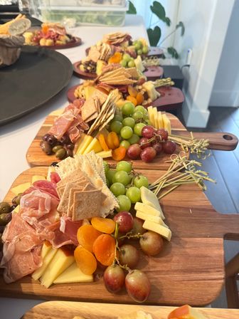 Colorful charcuterie boards lined up on a kitchen counter with green and red grapes, sliced cheeses, prosciutto and salami, assorted crackers, olives and dried apricots on wooden serving boards.