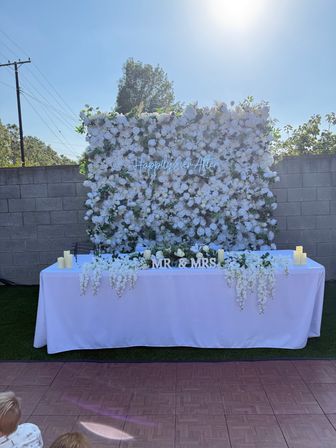 Outdoor backyard wedding head table with white tablecloth in front of a dense white rose floral wall, neon “Happily Ever After” sign, cascading white blooms, MR & MRS letters and battery candles under bright sun.