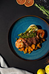 Gourmet surf-and-turf on a blue ceramic plate: sliced seared steak, grilled shrimp, chimichurri, diced sweet potatoes and charred baby bok choy, garnished with lemon and rosemary