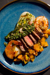 Vibrant surf-and-turf: seared medium-rare steak slices topped with chimichurri, grilled shrimp, diced sautéed squash and bok choy on a blue ceramic plate.