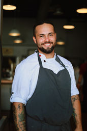 Smiling male chef in a white jacket and dark apron with tattooed forearms standing in a restaurant kitchen under pendant lights
