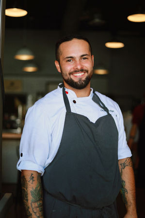 Smiling male chef in a white jacket and dark apron with tattooed forearms standing in a restaurant kitchen under pendant lights