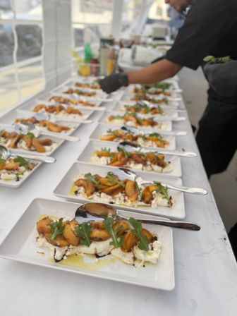 Rows of colorful plated appetizers — creamy burrata topped with roasted peaches, arugula, olive oil and balsamic glaze — lined up on a tented catering table as a server prepares them.