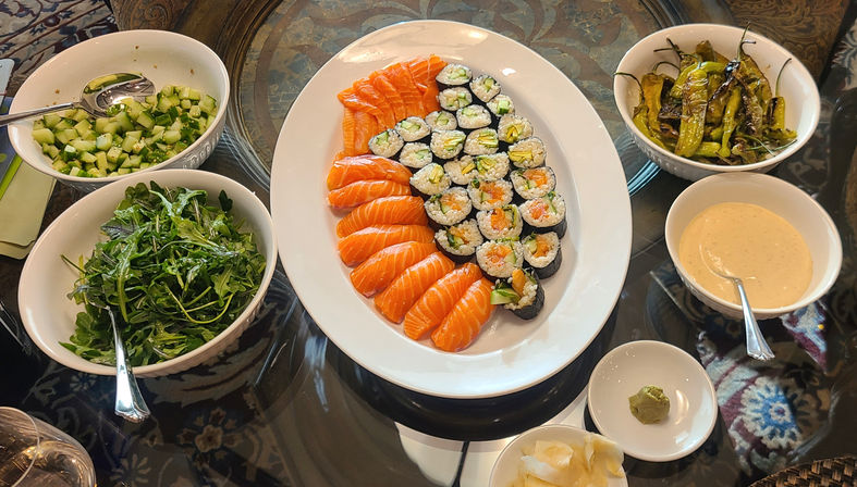 Colorful sushi platter with salmon nigiri and vegetable maki on a white oval plate, surrounded by bowls of arugula salad, diced cucumber, blistered shishito peppers, spicy mayo, wasabi and pickled ginger on a glass table.