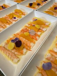 Rows of butterflied shrimp in white rectangular plates with orange segments, citrus dressing and colorful edible flower petals — bright seafood appetizer