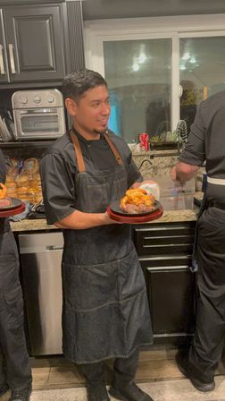Smiling chef in a dark denim apron holding a sizzling steak topped with shrimp on a wooden platter in a home kitchen with granite countertops and black cabinets