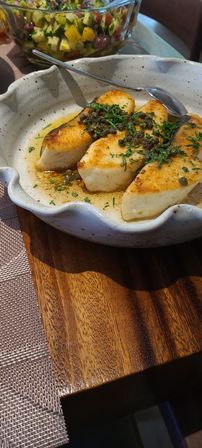 Seared white fish fillets topped with capers and chopped parsley in a rustic ceramic dish, spoon at the side, with a colorful cucumber‑tomato salsa in a glass bowl behind on a wooden table.