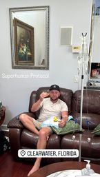 Relaxed man on a brown leather couch in Clearwater, Florida hooked to an IV drip while eating a sandwich, with a framed mirror, pillows and a 'Superhuman Protocol' text overlay.