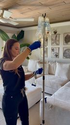 Medical clinician in blue gloves adjusting a saline IV bag on a pole during at-home IV therapy in a bright, coastal-style living room with beige sofa and botanical wall art