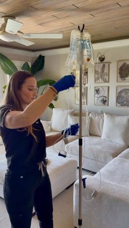 Home health professional in blue gloves adjusting an IV bag and tubing on a pole in a sunlit coastal-style living room with a white sectional sofa, tropical plant, and wood-paneled ceiling.