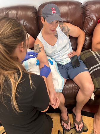 Phlebotomist in blue gloves performing an at-home blood draw on a tattooed man sitting on a brown leather couch wearing a tank top, baseball cap and flip-flops, phone resting on his thigh