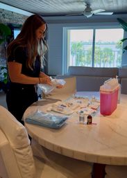 Person prepping sterile medical supplies, syringes and vials on a marble dining table in a bright waterfront living room with palm-tree lined balcony and water view