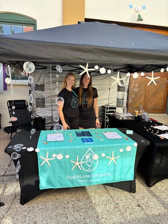 Two smiling women stand under a black canopy at an outdoor street-market wellness booth with a teal tablecloth, brochures, seashell and starfish decorations, and hanging bubble garlands.