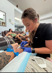 Healthcare professional in blue gloves performing an at-home blood draw on a tattooed arm in a casual living room while friends relax on a couch