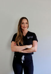 Confident healthcare professional in black scrubs with long wavy hair, smiling with arms crossed against a light gray wall — clean clinic portrait.
