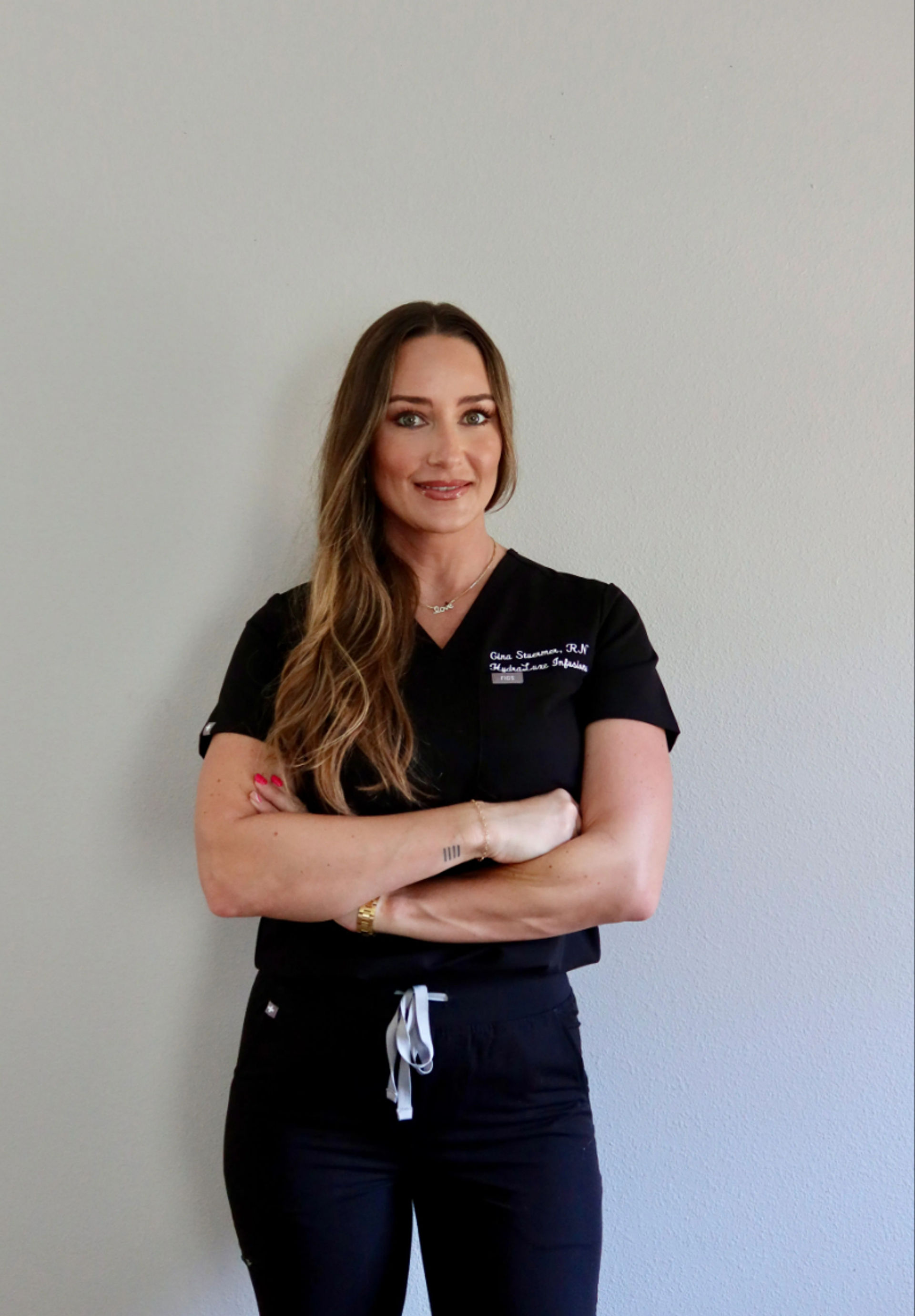 Confident healthcare professional in black scrubs, long wavy hair and arms crossed, smiling portrait against a light gray clinic wall